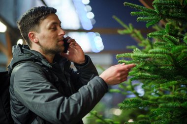 Şenlikli alışveriş, seçim ve Yeni Yıl nitelikleri satın alma. Beyaz adam telefonda konuşuyor ve yılbaşı arifesinde Noel ağacını seçmeyi tavsiye ediyor. Erkek ladin satın alır ve akıllı telefon kullanır..
