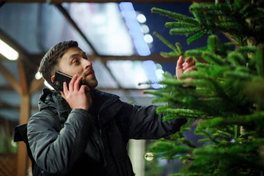 Şenlikli alışveriş, seçim ve Yeni Yıl nitelikleri satın alma. Beyaz adam telefonda konuşuyor ve yılbaşı arifesinde Noel ağacını seçmeyi tavsiye ediyor. Erkek ladin satın alır ve akıllı telefon kullanır..