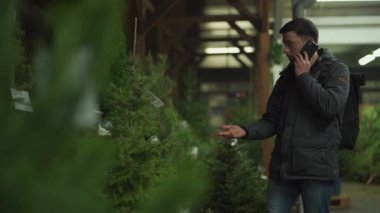 Şenlikli alışveriş, seçim ve Yeni Yıl nitelikleri satın alma. Beyaz adam telefonda konuşuyor ve yılbaşı arifesinde Noel ağacını seçmeyi tavsiye ediyor. Erkek ladin satın alır ve akıllı telefon kullanır.