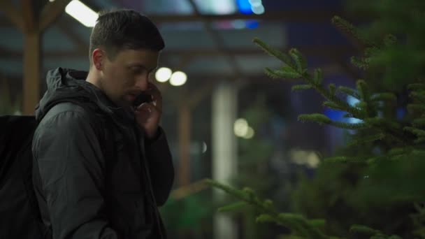 Homem caucasiano jovem escolhendo árvore de Natal em um mercado ao ar livre à noite falando por telefone com sua esposa e consultoria sobre a escolha certa. Símbolo de Natal e feriados de Ano Novo. Compras de férias — Vídeo de Stock