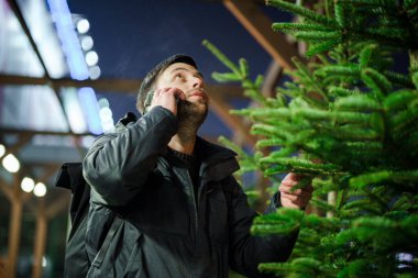 Şenlikli alışveriş, seçim ve Yeni Yıl nitelikleri satın alma. Beyaz adam telefonda konuşuyor ve yılbaşı arifesinde Noel ağacını seçmeyi tavsiye ediyor. Erkek ladin satın alır ve akıllı telefon kullanır..