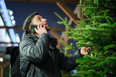 Şenlikli alışveriş, seçim ve Yeni Yıl nitelikleri satın alma. Beyaz adam telefonda konuşuyor ve yılbaşı arifesinde Noel ağacını seçmeyi tavsiye ediyor. Erkek ladin satın alır ve akıllı telefon kullanır..