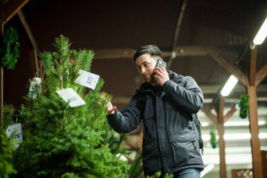 Şenlikli alışveriş, seçim ve Yeni Yıl nitelikleri satın alma. Beyaz adam telefonda konuşuyor ve yılbaşı arifesinde Noel ağacını seçmeyi tavsiye ediyor. Erkek ladin satın alır ve akıllı telefon kullanır..