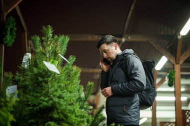 Şenlikli alışveriş, seçim ve Yeni Yıl nitelikleri satın alma. Beyaz adam telefonda konuşuyor ve yılbaşı arifesinde Noel ağacını seçmeyi tavsiye ediyor. Erkek ladin satın alır ve akıllı telefon kullanır..