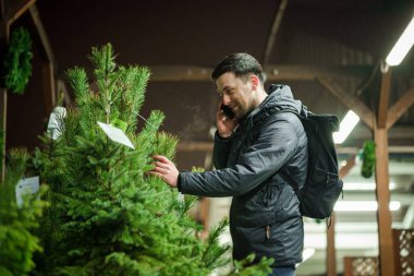 Şenlikli alışveriş, seçim ve Yeni Yıl nitelikleri satın alma. Beyaz adam telefonda konuşuyor ve yılbaşı arifesinde Noel ağacını seçmeyi tavsiye ediyor. Erkek ladin satın alır ve akıllı telefon kullanır..