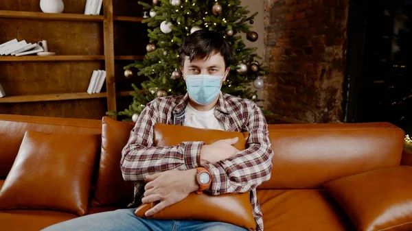 Depressed man in medical mask at home on the couch for Christmas. Coronavirus on Christmas Eve. New Year's holidays without friends and family during covid 19 lockdown and quarantine.