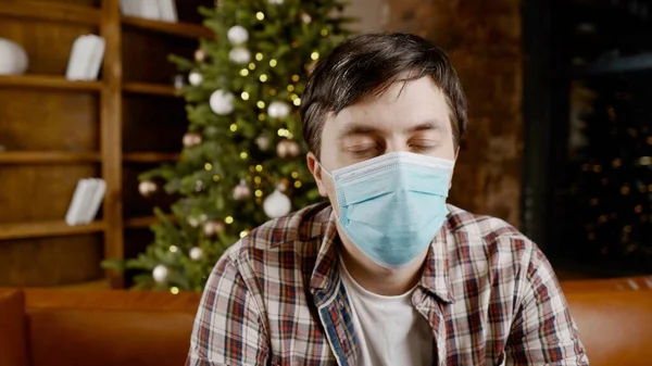 Depressed man in medical mask at home on the couch for Christmas. Coronavirus on Christmas Eve. New Year\'s holidays without friends and family during covid 19 lockdown and quarantine.