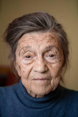 90 yaşında, gri saçlı, kırışık yüzlü, süveter giyen, portresi büyük, gülümseyen ve neşeli görünen yaşlı bir kadın. Tema: Karaciğer ve Yaşlanma, yaşlı insanların keyfi yerinde.
