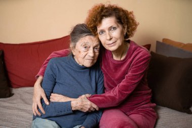 Anne ve kız, kızıl saçlı olgun bir kadın ve gri saçlı ve kırışıklıkları olan yaşlı, sevimli, tatlı bir kadın, evde kucaklaşın ve mutlu bir şekilde gülümseyin, aile bağları ve ebeveynleriyle ilgilenin..