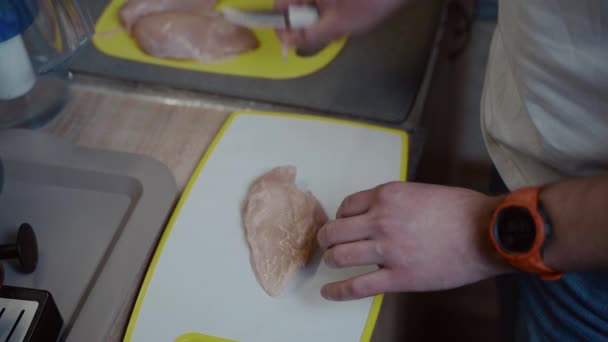 Le mani di primo piano di giovane maschio caucasico stanno tagliando la carne di pollo fresca su asse bianca di plastica a casa in cucina. Cucina da hobby, addio al celibato che prepara la cena. Un uomo solitario prepara il pollame per il pranzo — Video Stock