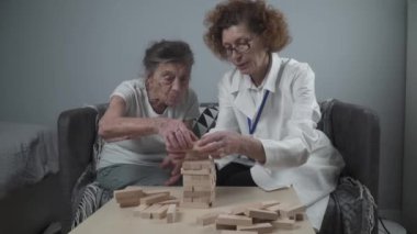 Jenga oyunu. Temaları bunama, yaşlanma ve yaşlılar için oyunlar. Kafkasyalı son sınıf öğrencisi kadın, hastaların evinde bir terapi ve oyun olarak bir doktorun yardımıyla ahşap bloklardan bir kule inşa ediyor.