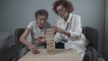 Jenga oyunu. Temaları bunama, yaşlanma ve yaşlılar için oyunlar. Kafkasyalı son sınıf öğrencisi kadın, bir hastalar evinde bir terapi ve jenga oyunu için bir doktorun yardımıyla ahşap bloklardan bir kule inşa ediyor.