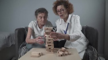 Jenga oyunu. Temaları bunama, yaşlanma ve yaşlılar için oyunlar. Kafkasyalı son sınıf öğrencisi kadın, hasta evinde bir terapi ve oyun için bir doktorun yardımıyla ahşap bloklardan bir kule inşa ediyor.