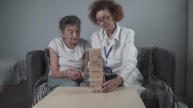 Jenga oynayan yaşlı kadın, bloklardan kule inşa ediyor. Beyaz laboratuvar önlüklü yaşlı doktor, kıdemli hastayı destekliyor, mantık geliştiriyor, iyi motor beceriler geliştiriyor, evde yaşlı yalnız kadın hastalıkları var.