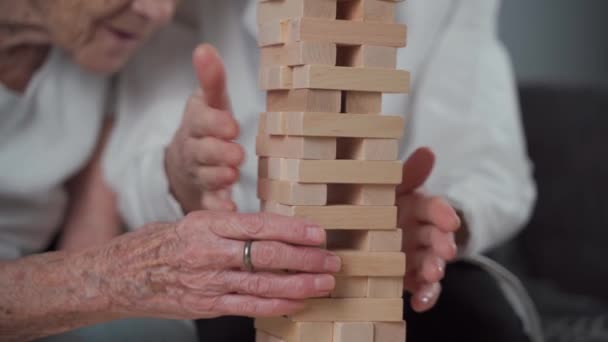 Старша жінка практикує навички будівництва дерев'яних блоків, будівництва вежі і намагається не допустити падіння, гра. Старий пацієнт витягує блок, розміщує зверху, підтримує топорею під час терапевтичної деменції в будинку — стокове відео