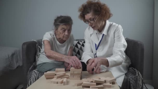 Senior Frau Jenga spielen, bauen Turm aus Blöcken. Älterer Arzt im weißen Laborkittel, unterstützt ältere Patientin, entwickelt Logik, Feinmotorik, Alzheimer zu Hause bei alten einsamen Frauen — Stockvideo