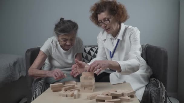 Умалишённая терапия игриво, тренировка пальцев и мелкая моторика, постройка деревянных блоков в башню, игра в Дженгу. Старшая женщина 90 лет и врач играют в образовательные игры в доме престарелых — стоковое видео