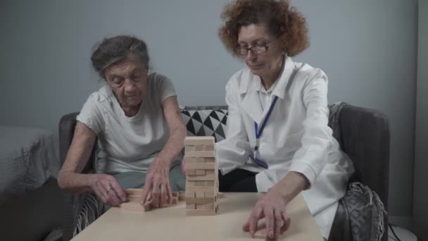 Senior Frau Jenga spielen, bauen Turm aus Blöcken. Älterer Arzt im weißen Laborkittel, unterstützt ältere Patientin, entwickelt Logik, Feinmotorik, Alzheimer zu Hause bei alten einsamen Frauen — Stockvideo