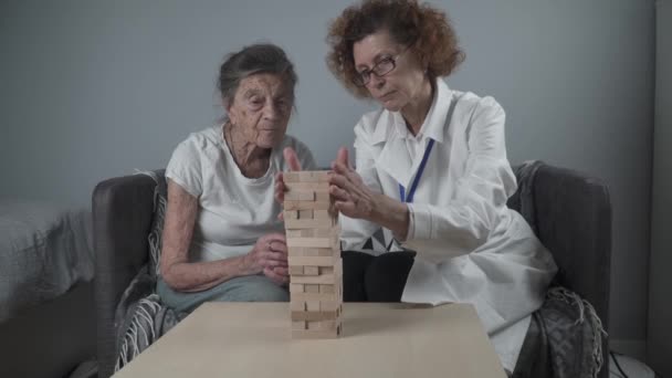 As habilidades de prática da mulher sênior constroem blocos de madeira, construindo torre e tentando não deixá-la cair, jogo de Jenga. Paciente idoso retira bloco, coloca em cima, suporte dotor durante a terapia demência em casa — Vídeo de Stock