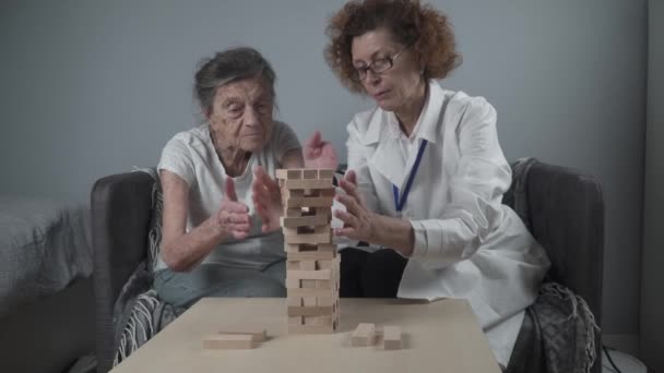 Starsza kobieta grająca Jengę, buduje wieżę z klocków. Starszy lekarz w białym fartuchu laboratoryjnym, wspomagający starszego pacjenta, rozwijający logikę, zdolności motoryczne, choroba Alzheimera w domu ze starą samotną kobietą — Wideo stockowe