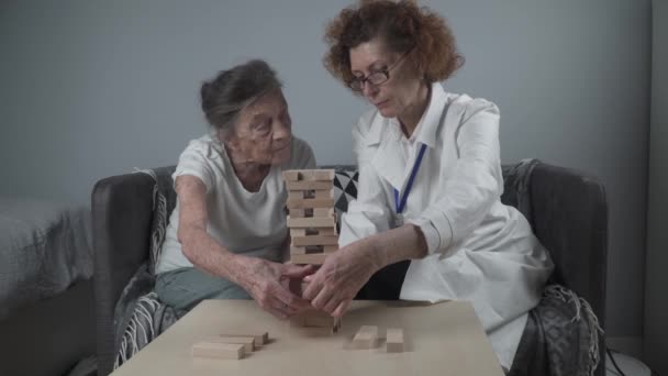 Jengaゲーム。テーマは高齢者の認知症、高齢化、ゲームです。白人のシニア女性は、患者の家で治療とjengaゲームの一環として医師の助けを借りて木製のブロックの塔を構築します。 — ストック動画