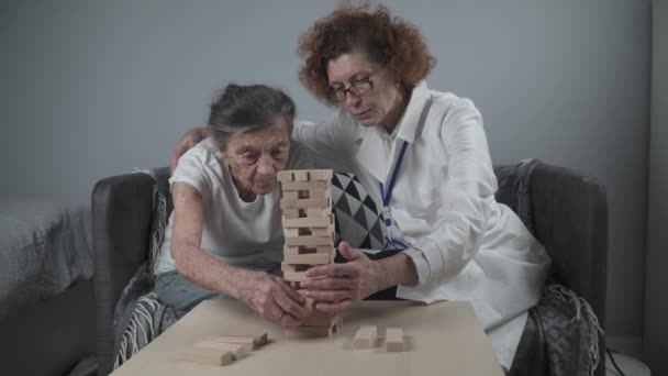 上級女性の練習のスキルは、木製のブロックを構築し、タワーを構築し、それを秋にしないようにしようとすると、 Jengaゲーム。古い患者は、ブロックを引き出します,上の場所,家の中で治療認知症の間にドアをサポート — ストック動画