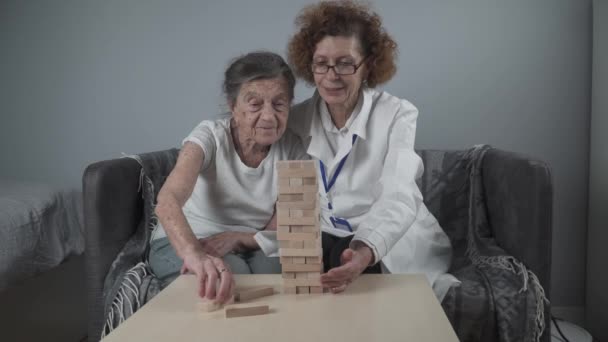 Jenga meccs. Téma a demencia, öregedés és játékok az idősek számára. Kaukázusi idős nő épít torony fa blokkok segítségével egy orvos részeként a terápia és a jenga játék egy beteg otthon — Stock videók