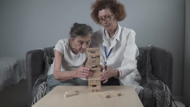 Terapia de demencia de manera lúdica, entrenamiento de dedos y habilidades motoras finas, construir bloques de madera en la torre, jugando Jenga. Mujer mayor de 90 años y médico jugando juego educativo en el hogar de ancianos — Vídeos de Stock