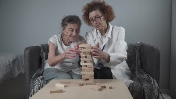 Старшая женщина практиковать навыки построить деревянные блоки, строительство башни и стараясь не дать ему упасть, игра Дженга. Старый пациент вынимает блок, место на вершине, поддержка врача во время терапии слабоумия в доме — стоковое видео