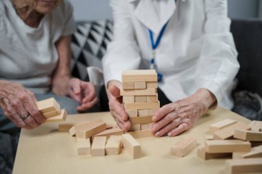 Jenga oyunu. Temaları bunama, yaşlanma ve yaşlılar için oyunlar. Kafkasyalı son sınıf öğrencisi kadın, bir hastanın evindeki terapi ve jenga oyununun bir parçası olarak bir doktorun yardımıyla ahşap bloklardan kule inşa ediyor..