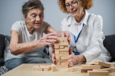 Jenga oyunu. Temaları bunama, yaşlanma ve yaşlılar için oyunlar. Kafkasyalı son sınıf öğrencisi kadın, bir hastanın evindeki terapi ve jenga oyununun bir parçası olarak bir doktorun yardımıyla ahşap bloklardan kule inşa ediyor..