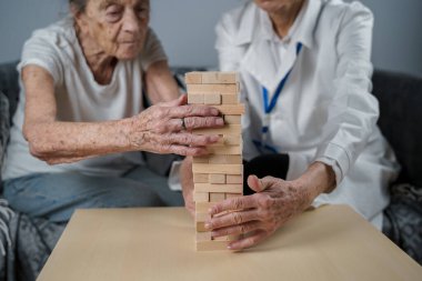 Jenga oyunu. Temaları bunama, yaşlanma ve yaşlılar için oyunlar. Kafkasyalı son sınıf öğrencisi kadın, bir hastanın evindeki terapi ve jenga oyununun bir parçası olarak bir doktorun yardımıyla ahşap bloklardan kule inşa ediyor..