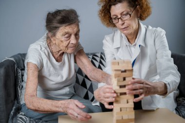 Jenga oynayan yaşlı kadın, bloklardan kule inşa ediyor. Beyaz laboratuvar önlüklü yaşlı doktor, kıdemli hastayı destekliyor, mantık geliştiriyor, iyi motor beceriler geliştiriyor, evde yaşlı yalnız kadın hastalıkları var..