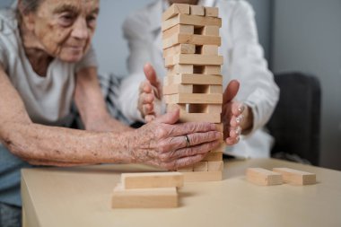 Jenga oyunu. Temaları bunama, yaşlanma ve yaşlılar için oyunlar. Kafkasyalı son sınıf öğrencisi kadın, bir hastanın evindeki terapi ve jenga oyununun bir parçası olarak bir doktorun yardımıyla ahşap bloklardan kule inşa ediyor..