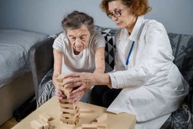 Jenga oynayan yaşlı kadın, bloklardan kule inşa ediyor. Beyaz laboratuvar önlüklü yaşlı doktor, kıdemli hastayı destekliyor, mantık geliştiriyor, iyi motor beceriler geliştiriyor, evde yaşlı yalnız kadın hastalıkları var..