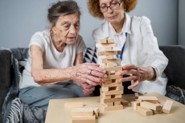 Jenga oyunu. Temaları bunama, yaşlanma ve yaşlılar için oyunlar. Kafkasyalı son sınıf öğrencisi kadın, bir hastanın evindeki terapi ve jenga oyununun bir parçası olarak bir doktorun yardımıyla ahşap bloklardan kule inşa ediyor..