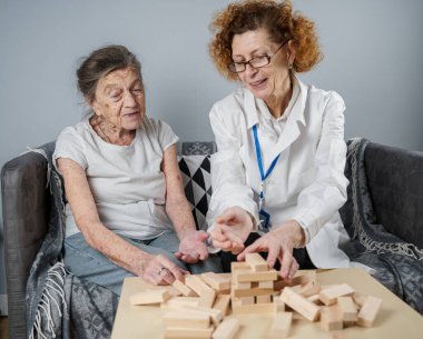 Jenga oyunu. Temaları bunama, yaşlanma ve yaşlılar için oyunlar. Kafkasyalı son sınıf öğrencisi kadın, bir hastanın evindeki terapi ve jenga oyununun bir parçası olarak bir doktorun yardımıyla ahşap bloklardan kule inşa ediyor..