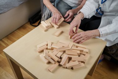 Jenga oyunu. Temaları bunama, yaşlanma ve yaşlılar için oyunlar. Kafkasyalı son sınıf öğrencisi kadın, bir hastanın evindeki terapi ve jenga oyununun bir parçası olarak bir doktorun yardımıyla ahşap bloklardan kule inşa ediyor..