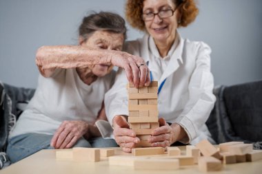Jenga oyunu. Temaları bunama, yaşlanma ve yaşlılar için oyunlar. Kafkasyalı son sınıf öğrencisi kadın, bir hastanın evindeki terapi ve jenga oyununun bir parçası olarak bir doktorun yardımıyla ahşap bloklardan kule inşa ediyor..