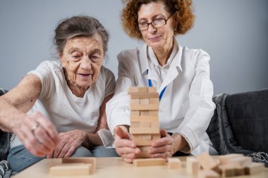 Jenga oyunu. Temaları bunama, yaşlanma ve yaşlılar için oyunlar. Kafkasyalı son sınıf öğrencisi kadın, bir hastanın evindeki terapi ve jenga oyununun bir parçası olarak bir doktorun yardımıyla ahşap bloklardan kule inşa ediyor..