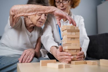 Jenga oyunu. Temaları bunama, yaşlanma ve yaşlılar için oyunlar. Kafkasyalı son sınıf öğrencisi kadın, bir hastanın evindeki terapi ve jenga oyununun bir parçası olarak bir doktorun yardımıyla ahşap bloklardan kule inşa ediyor..