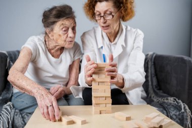 Jenga oyunu. Temaları bunama, yaşlanma ve yaşlılar için oyunlar. Kafkasyalı son sınıf öğrencisi kadın, bir hastanın evindeki terapi ve jenga oyununun bir parçası olarak bir doktorun yardımıyla ahşap bloklardan kule inşa ediyor..