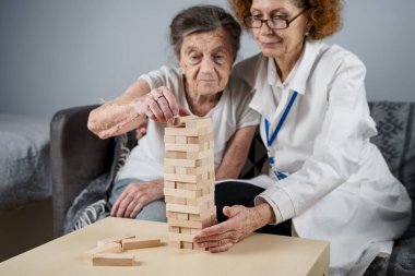 Jenga oyunu. Temaları bunama, yaşlanma ve yaşlılar için oyunlar. Kafkasyalı son sınıf öğrencisi kadın, bir hastanın evindeki terapi ve jenga oyununun bir parçası olarak bir doktorun yardımıyla ahşap bloklardan kule inşa ediyor..