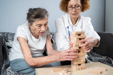 Jenga oyunu. Temaları bunama, yaşlanma ve yaşlılar için oyunlar. Kafkasyalı son sınıf öğrencisi kadın, bir hastanın evindeki terapi ve jenga oyununun bir parçası olarak bir doktorun yardımıyla ahşap bloklardan kule inşa ediyor..