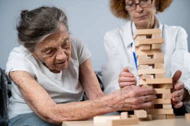 Jenga oyunu. Temaları bunama, yaşlanma ve yaşlılar için oyunlar. Kafkasyalı son sınıf öğrencisi kadın, bir hastanın evindeki terapi ve jenga oyununun bir parçası olarak bir doktorun yardımıyla ahşap bloklardan kule inşa ediyor..