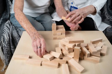 Jenga oyunu. Temaları bunama, yaşlanma ve yaşlılar için oyunlar. Kafkasyalı son sınıf öğrencisi kadın, bir hastanın evindeki terapi ve jenga oyununun bir parçası olarak bir doktorun yardımıyla ahşap bloklardan kule inşa ediyor..