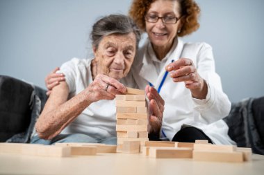Jenga oyunu. Temaları bunama, yaşlanma ve yaşlılar için oyunlar. Kafkasyalı son sınıf öğrencisi kadın, bir hastanın evindeki terapi ve jenga oyununun bir parçası olarak bir doktorun yardımıyla ahşap bloklardan kule inşa ediyor..