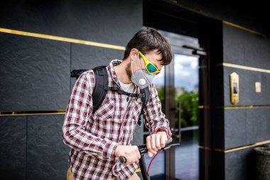 Ön kapıda müşteri bekleyen Kafkasyalı genç kurye, elektrikli scooter ile geldi, sis ve toza karşı koruyucu maske takarak saatine baktı. Çevre dostu ulaşım ve teslimat.