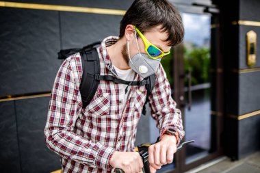 Kurye şehirde elektrikli scooter kullanıyor. Sırt çantalı, şık giyinmiş, şehirde hava kirliliğine karşı maske takan, müşteri bekleyen ve kol saatine bakan çekici beyaz bir adam..