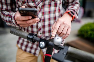 Erkek eller modern elektrikli scooter çalışır ve akort eder. Kafkasyalı bir adam şık giyinmiş. Mobiletini kontrol etmek için cep telefonu kullanıyor. Yüzü görünmüyor. Kentte kişisel elektrik taşımacılığı.